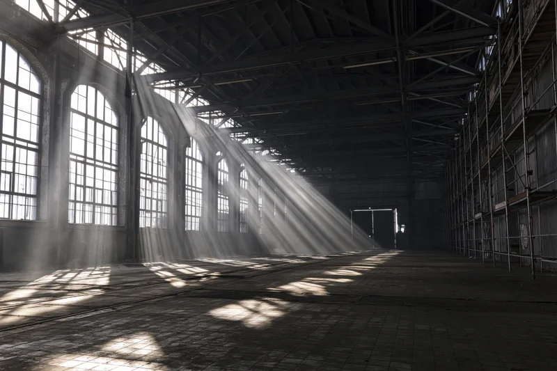 Empty industrial warehouse interior with sunlight streaming through arched windows onto concrete floor