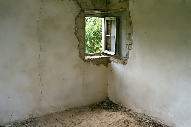 Corner of an empty abandoned room with cracked plaster walls and a broken window opening to green foliage