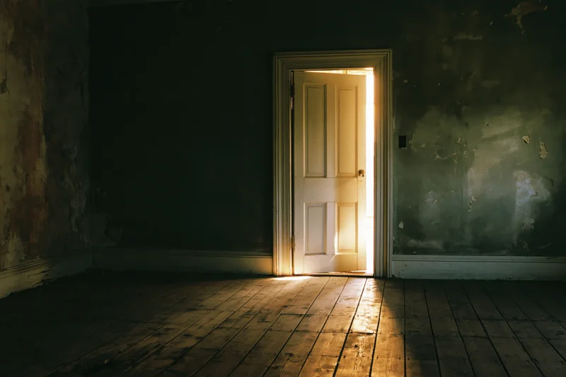 Ajar white door in an empty abandoned room letting in warm golden light onto wooden floor
