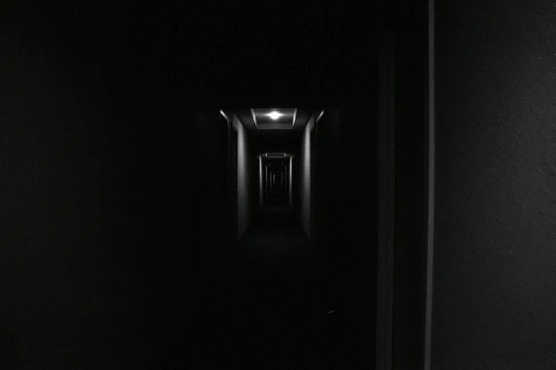 Dark hotel corridor receding into near-total darkness with a single ceiling light at the end