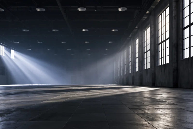 Empty industrial warehouse interior with light rays through tall windows on concrete floor