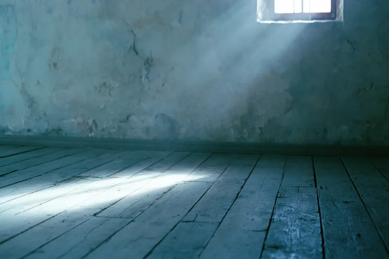 Empty room with weathered wooden floorboards and a barred window letting in a beam of light