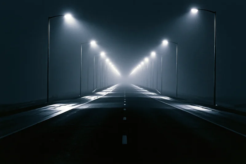Empty two-lane highway at night illuminated by streetlights receding into fog