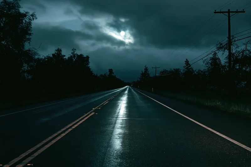 Wet two-lane highway stretching to vanishing point under moonlit cloudy sky at night