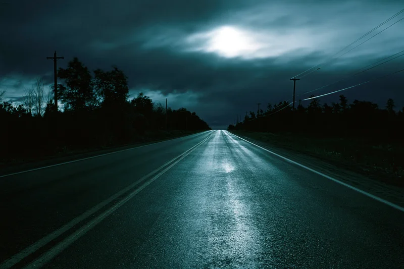 Empty two-lane highway stretching to vanishing point under moonlit stormy sky