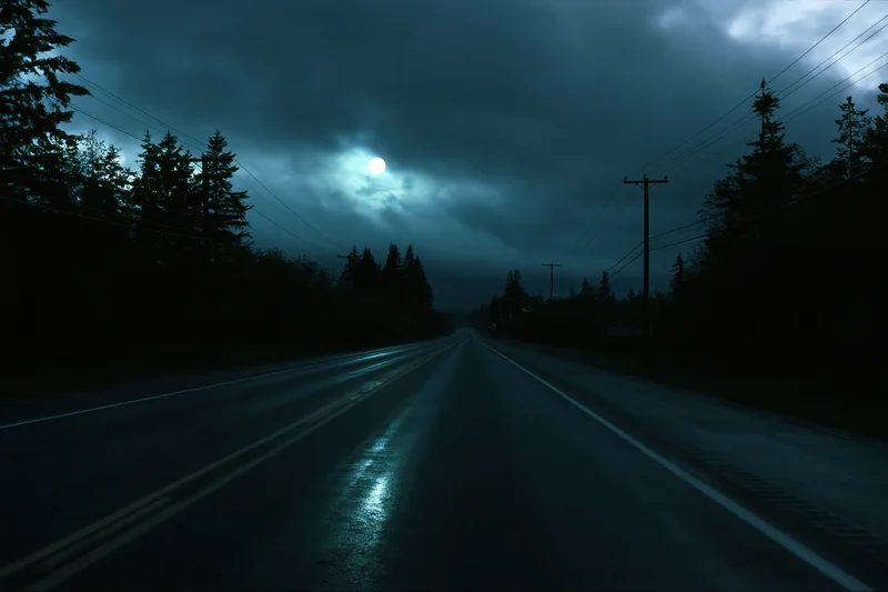 Wet two-lane highway stretching into the distance under a cloudy moonlit sky at night