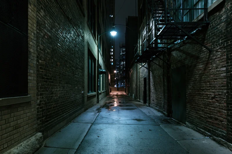 Narrow alley between old brick warehouse buildings at night, lit by a single lamppost, wet pavement reflecting light