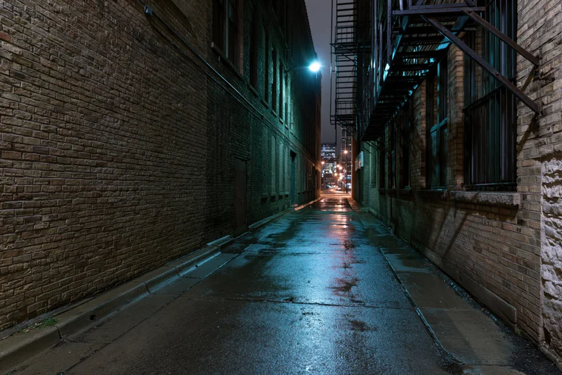 Narrow wet alley between old brick warehouse buildings at night with blue streetlight reflections on pavement