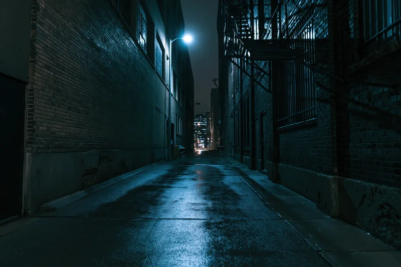 Narrow alley between old brick warehouse buildings at night, illuminated by a single lamppost, wet pavement reflecting light