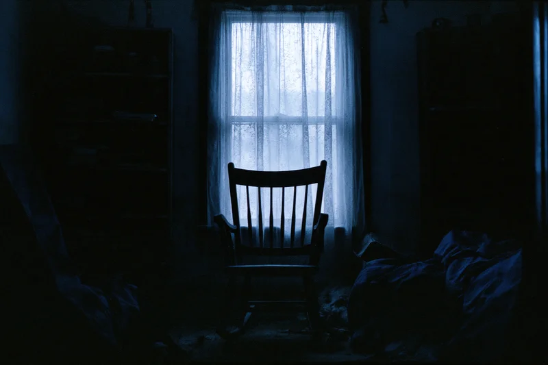 Wooden rocking chair silhouetted in a dark room by a curtained window