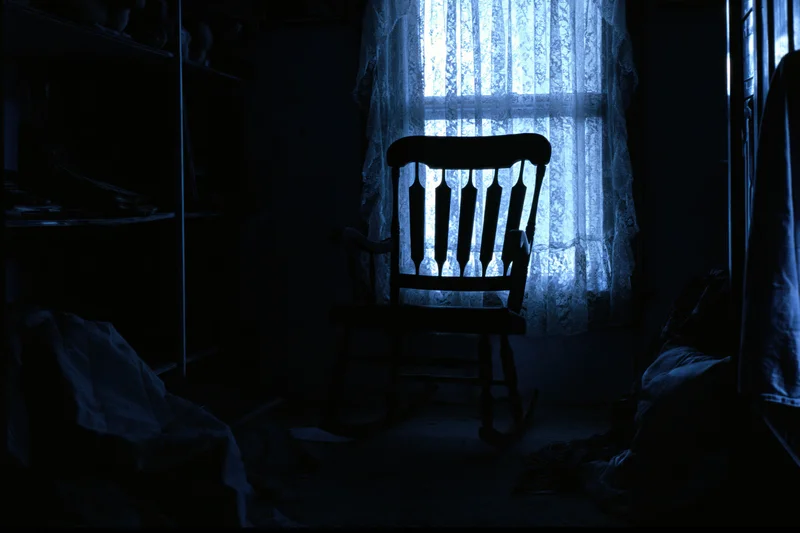 Wooden rocking chair silhouetted in a dark room by light through lace curtain