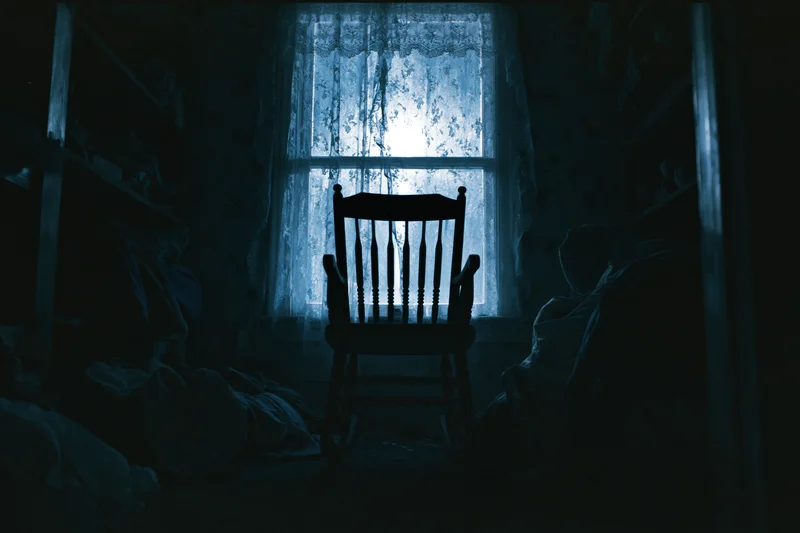 Wooden rocking chair silhouetted in dark room centered before window with lace curtain