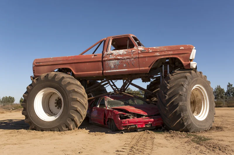 Rust-red monster truck crushing a red car on a dirt field under clear blue sky