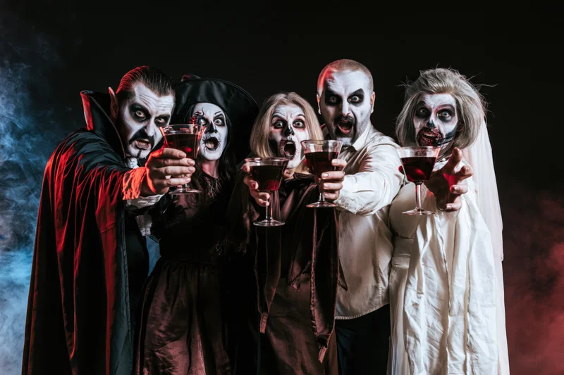 Five adults in vampire costumes holding cocktail glasses in a smoky dark setting for a halloween party