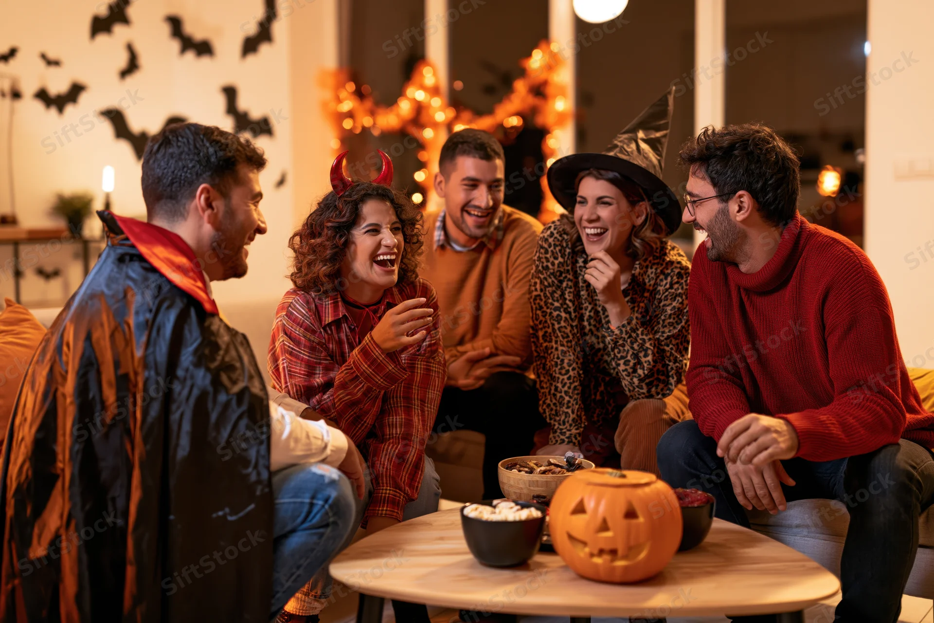 Five adults in halloween costumes laughing and chatting around a table in a decorated living room