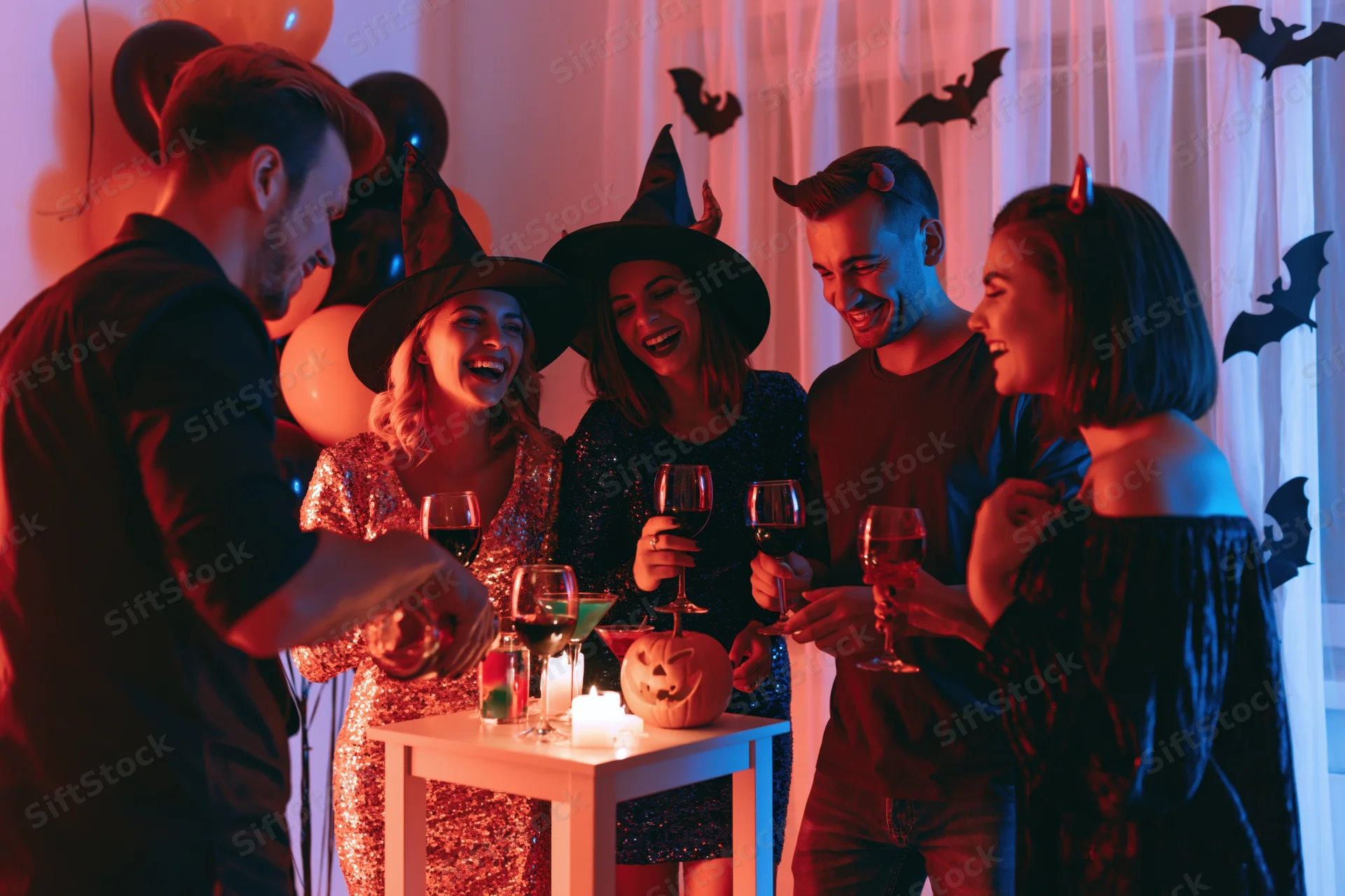 Five friends in halloween costumes toasting with wine around a decorated table indoors