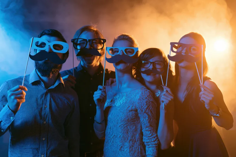 Five young adults posing with paper mustache and glasses props at a smoky nightclub party