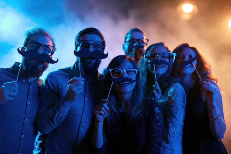 Young adults smiling with paper mustache and glasses props at a smoky nightclub party