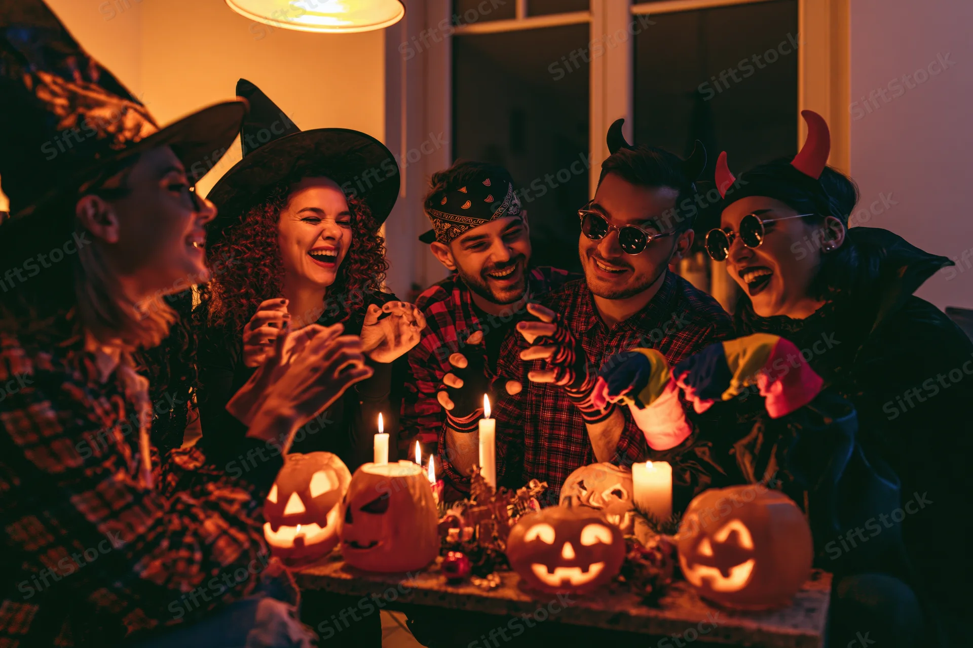 Five young adults in halloween costumes laughing around a table with lit pumpkins and candles indoors