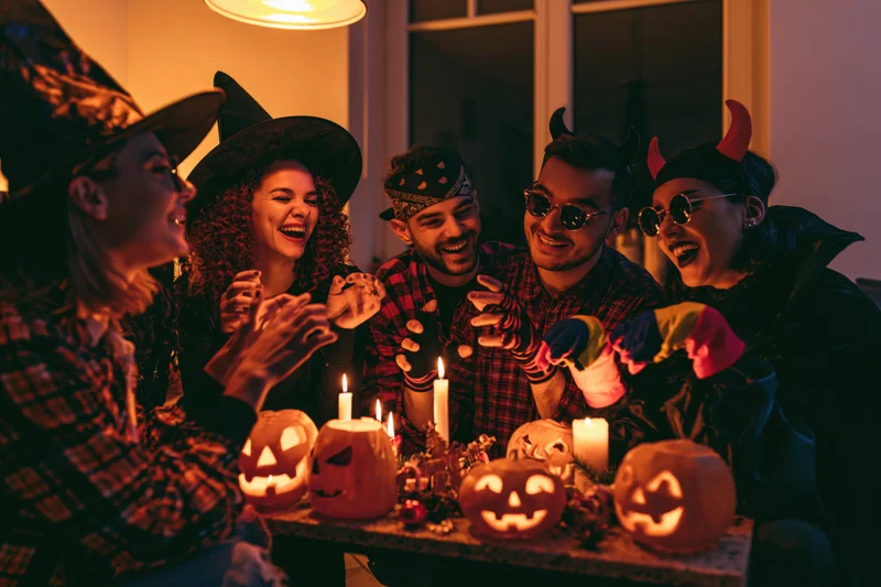 Five young adults in halloween costumes laughing around a table with lit pumpkins and candles indoors