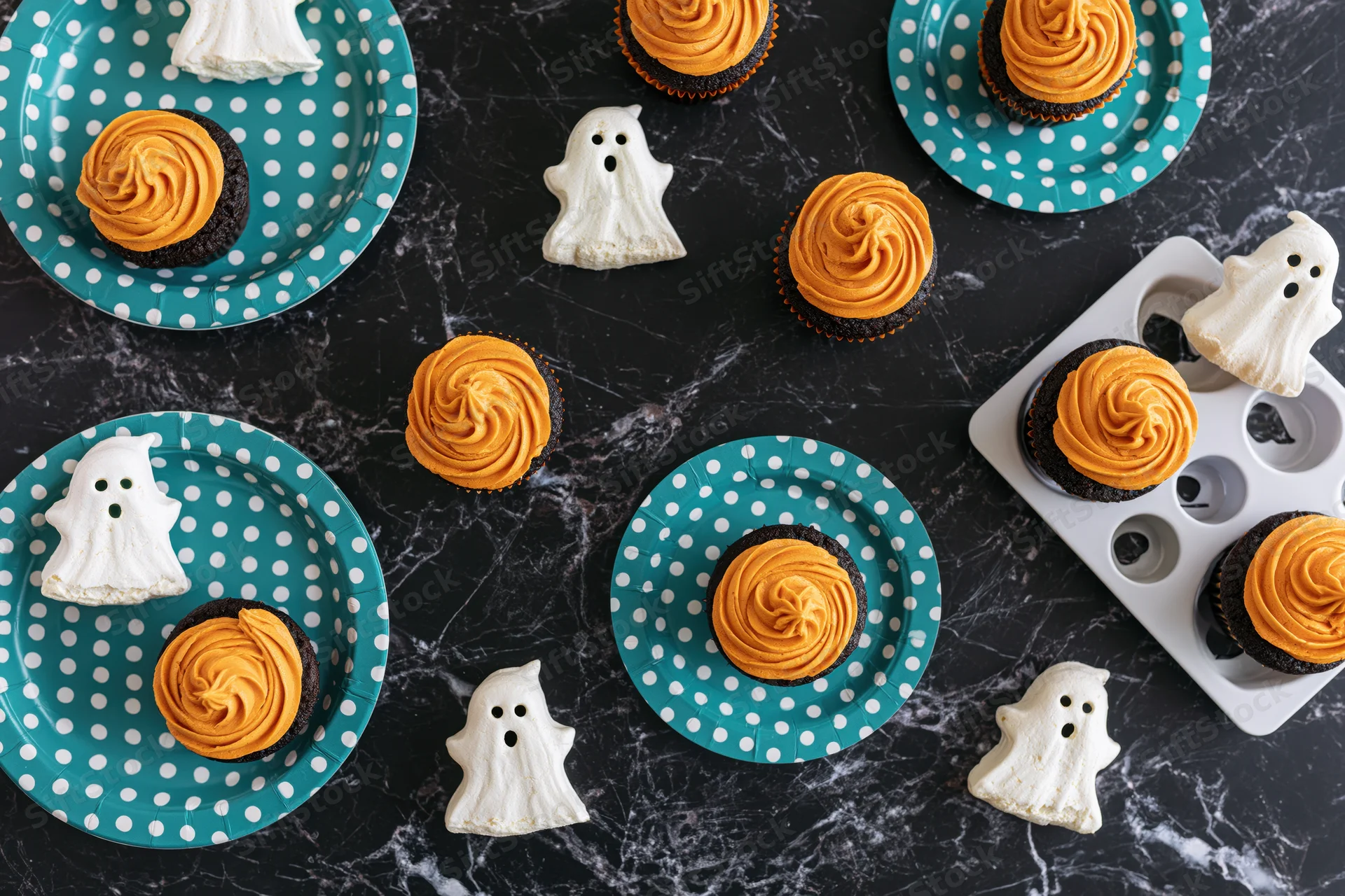 Halloween cupcakes with orange frosting and ghost cookies arranged on teal polka dot plates and dark marble surface