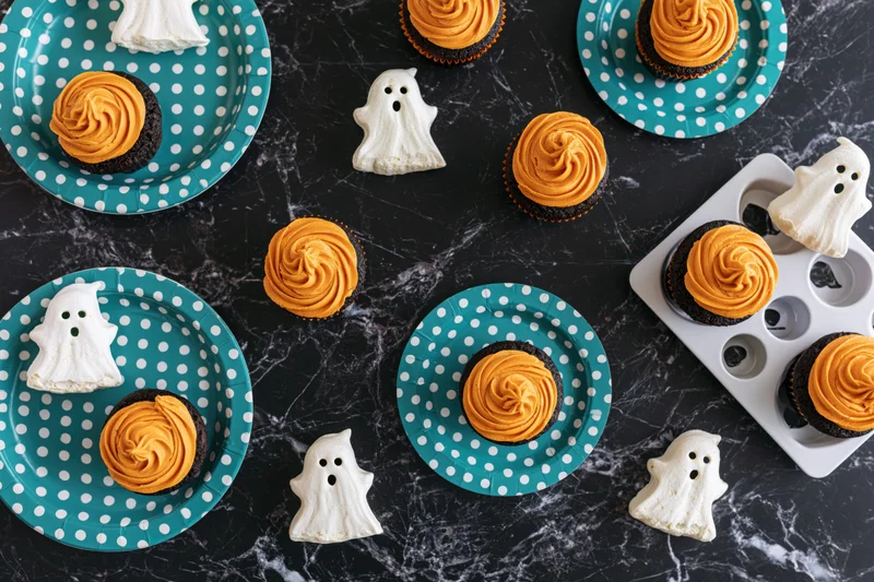 Halloween cupcakes with orange frosting and ghost cookies arranged on teal polka dot plates and dark marble surface