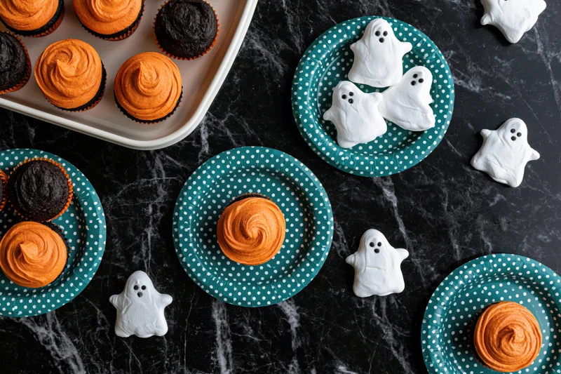 Halloween-themed cupcakes with ghost-shaped meringues on teal polka dot plates arranged on a black marble surface