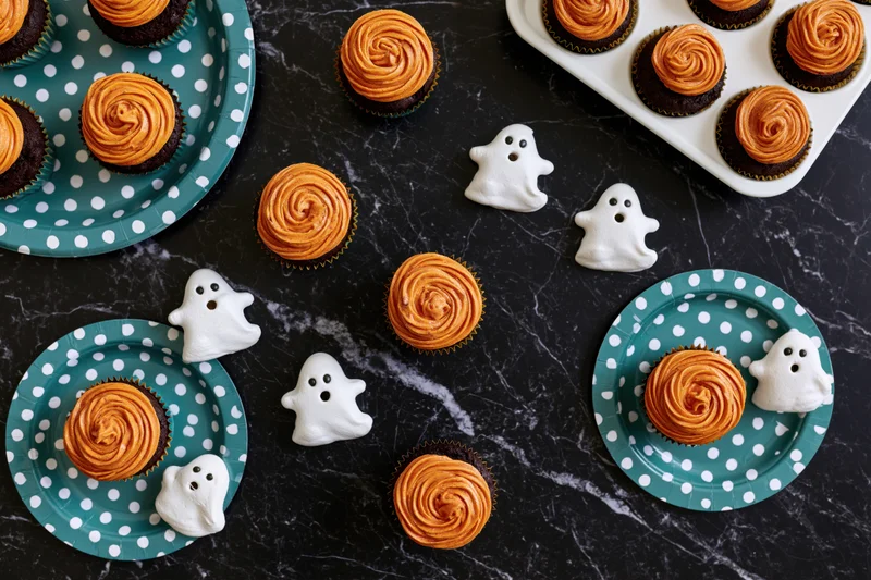 Halloween-themed cupcakes with orange frosting and white ghost candies arranged on dark marble surface with teal polka dot plates