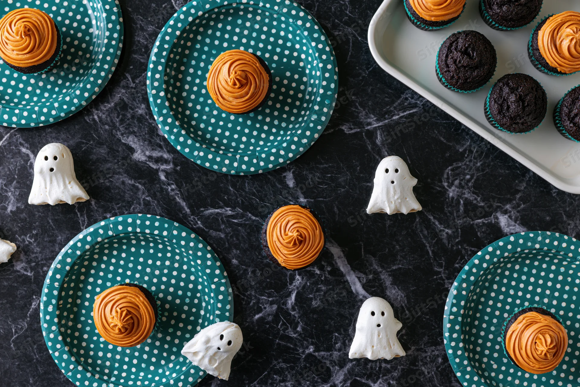 Halloween cupcakes with orange frosting and white ghost candies arranged on teal polka dot plates and dark marble surface