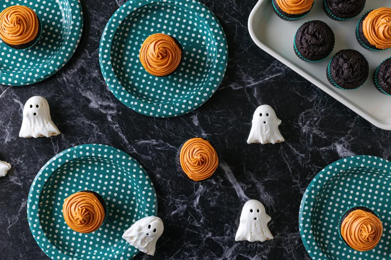 Halloween cupcakes with orange frosting and white ghost candies arranged on teal polka dot plates and dark marble surface