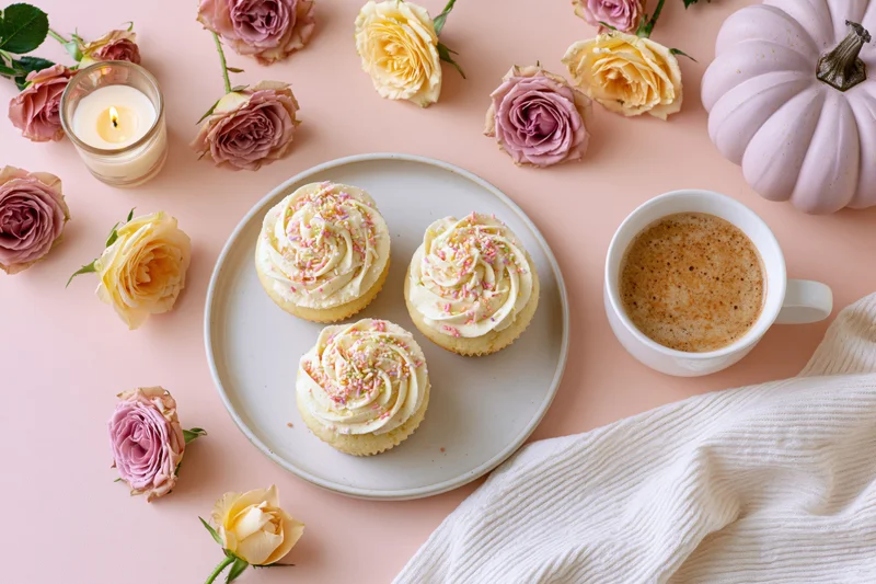 Three frosted cupcakes with pastel sprinkles on a plate surrounded by roses, a pumpkin, coffee, and a candle on a pink surface