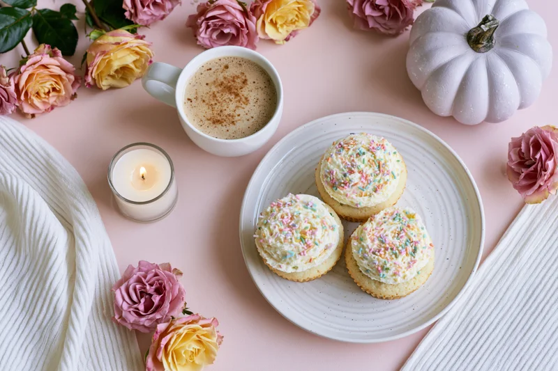 Three frosted cupcakes with pastel sprinkles on a white plate surrounded by roses, a pumpkin, a candle, and a coffee mug on a pink surface