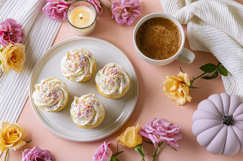 Four frosted cupcakes with pastel sprinkles on a white plate surrounded by roses, a coffee mug, a candle, and a purple pumpkin on a pink surface