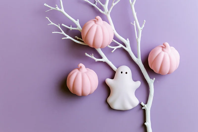 Flat lay of pink ceramic pumpkins and white ghost ornament on purple background with white branch