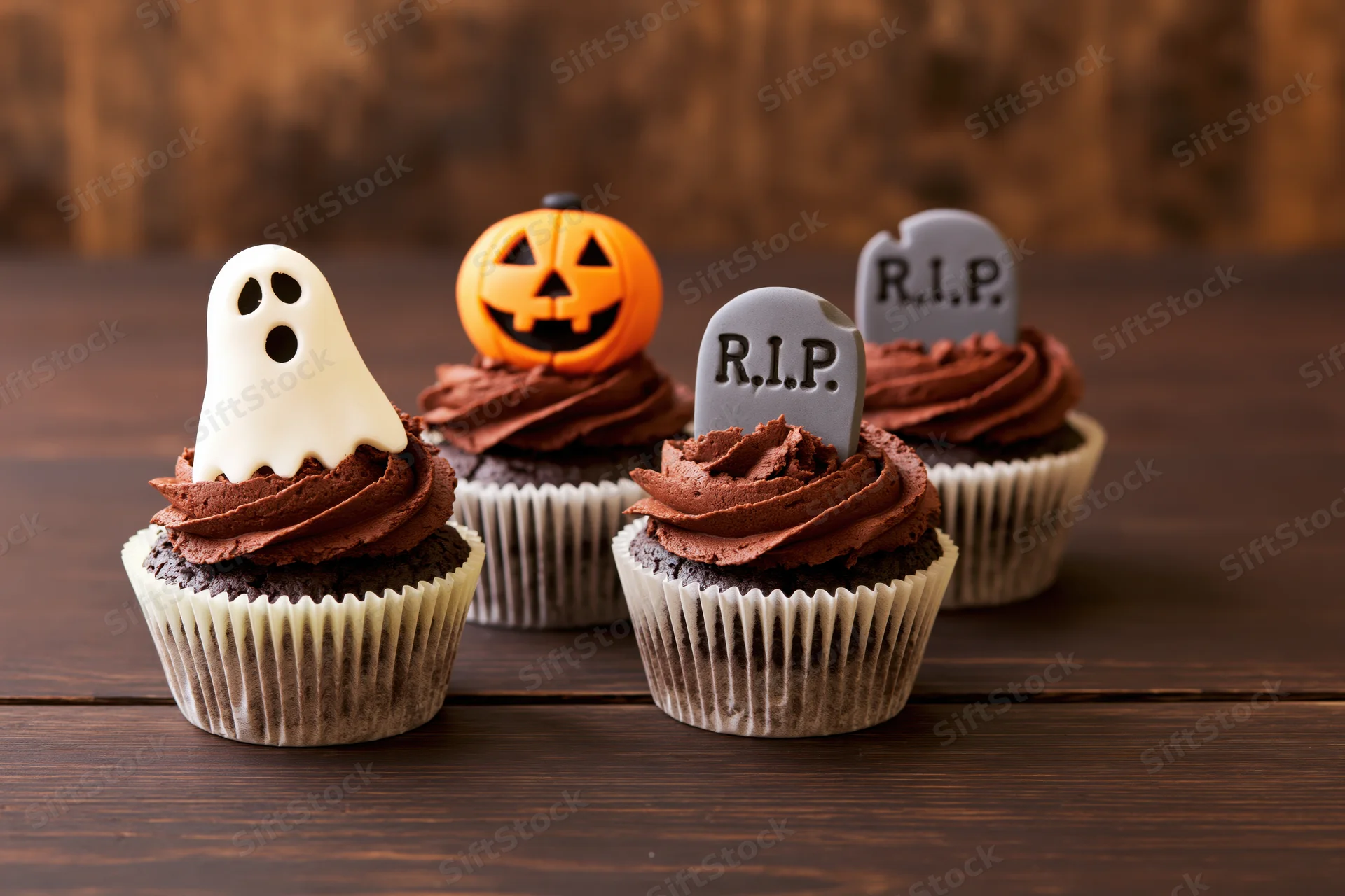 Four halloween-themed cupcakes with ghost, pumpkin, and tombstone decorations on a dark wooden table
