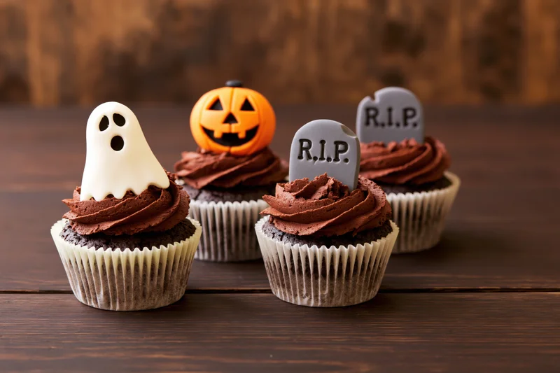 Four halloween-themed cupcakes with ghost, pumpkin, and tombstone decorations on a dark wooden table