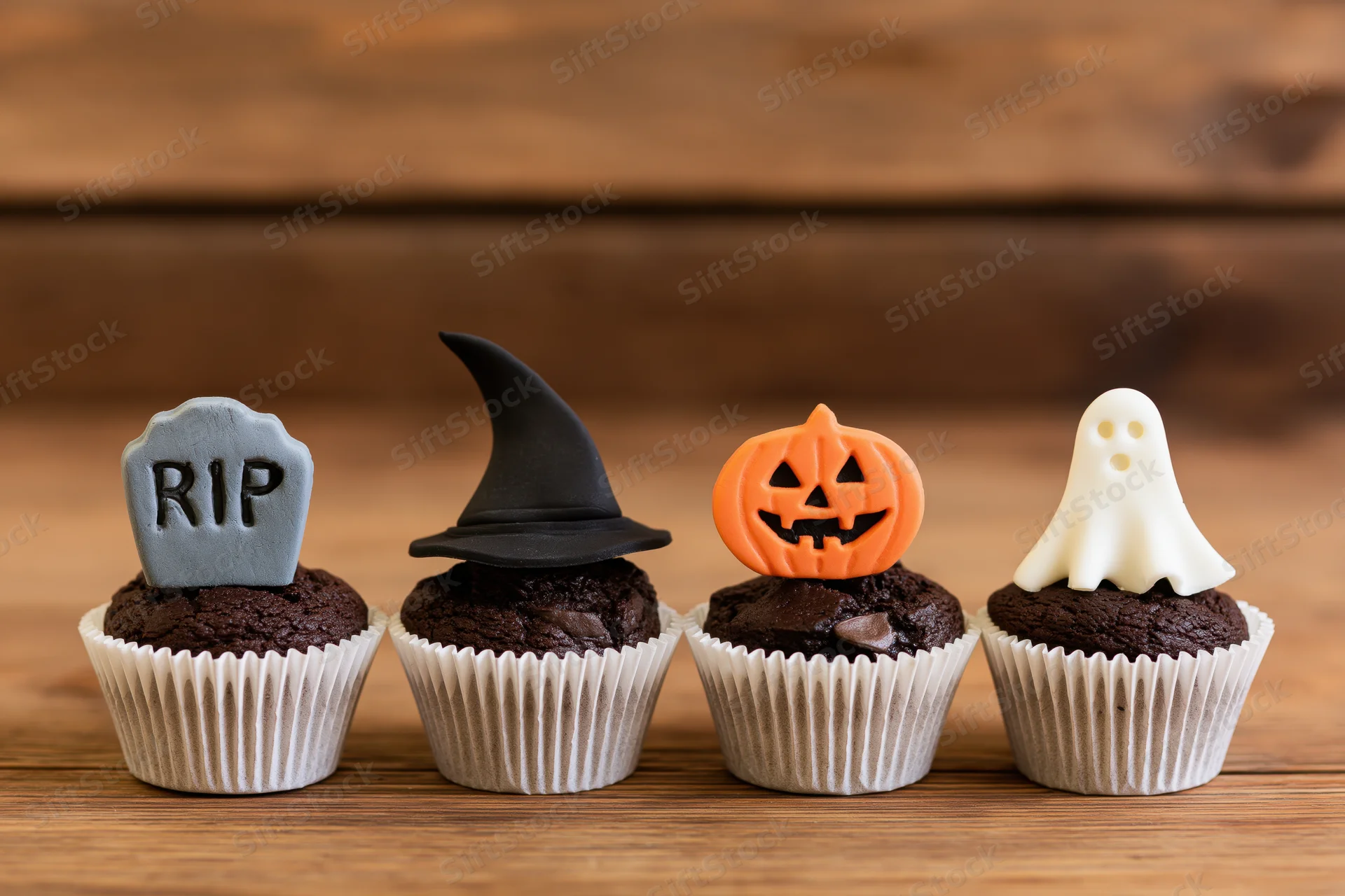 Four halloween chocolate cupcakes with tombstone, witch hat, jack-o-lantern and ghost decorations on wooden table