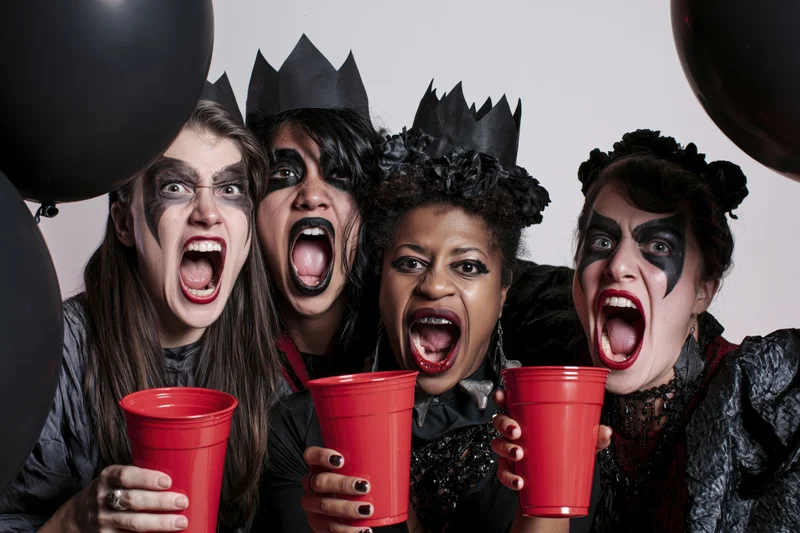 Four women in halloween costumes with theatrical makeup screaming while holding red cups and black balloons in a studio setting