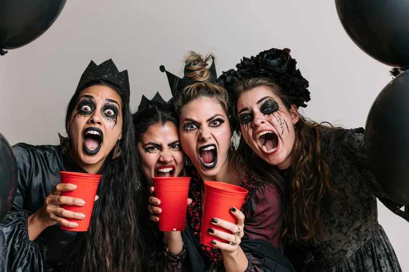 Four women in halloween costumes with theatrical makeup screaming while holding red cups at a party