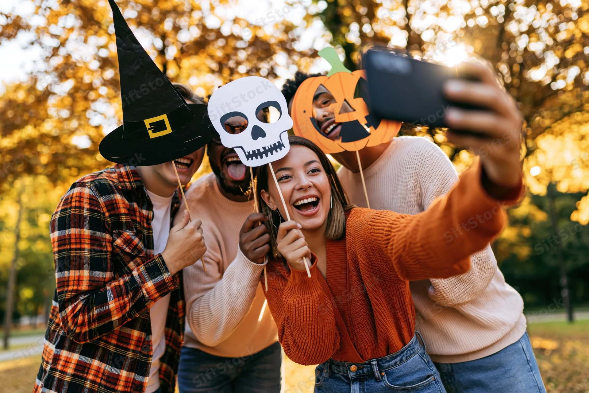 Four friends taking a group selfie outdoors with halloween props in a park during autumn