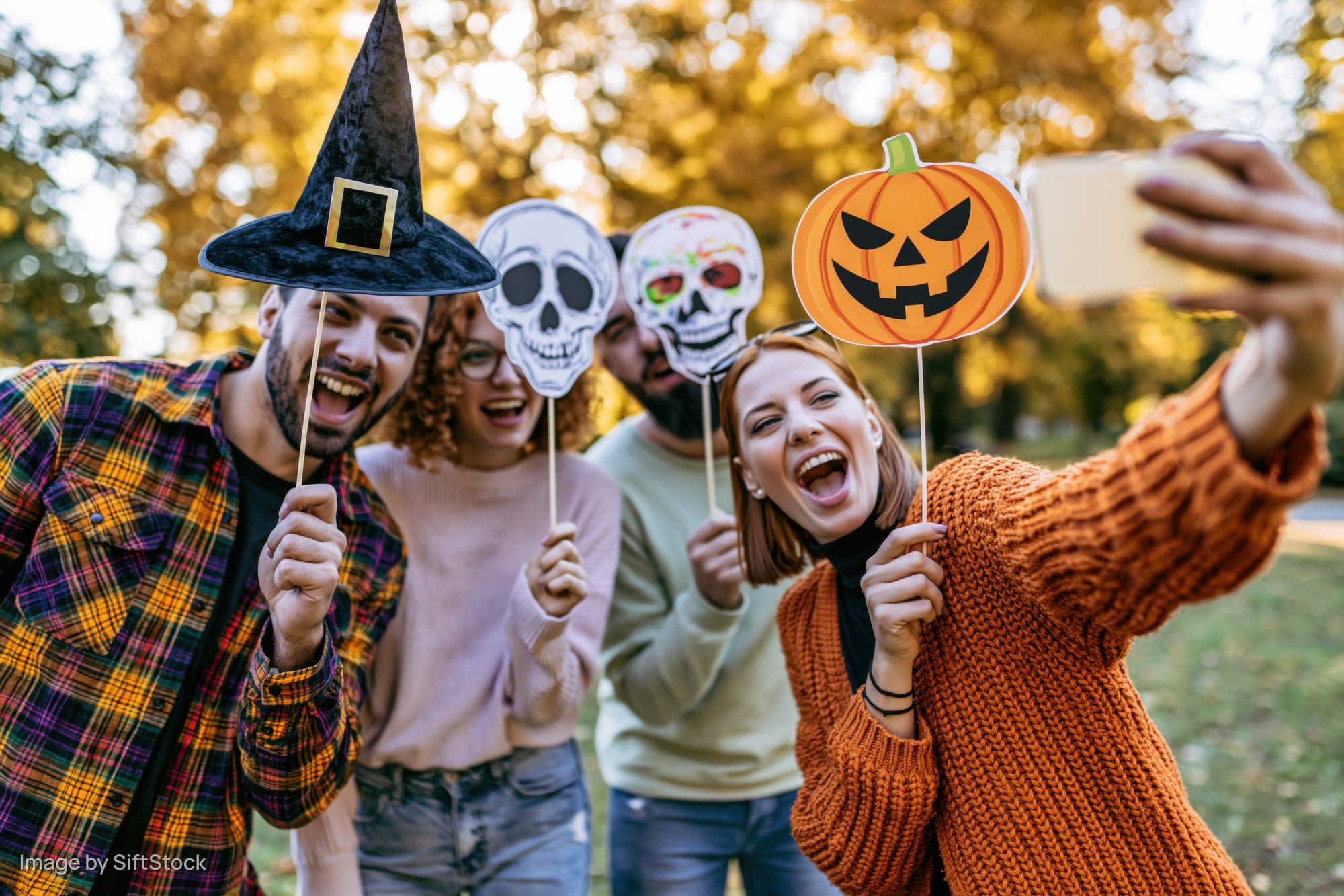 Four friends taking a group selfie outdoors with halloween props in a park during autumn