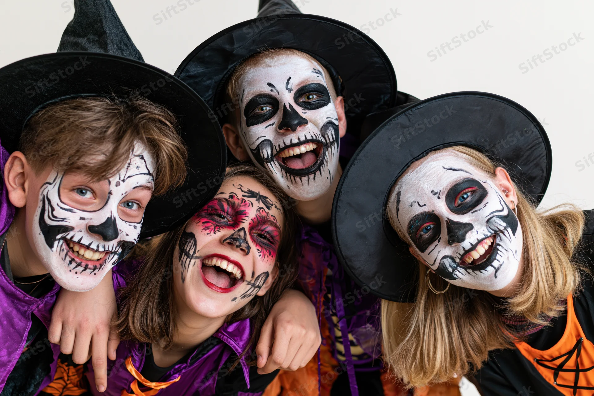 Four teenagers in halloween costumes with skull face paint and witch hats smiling together in a group pose
