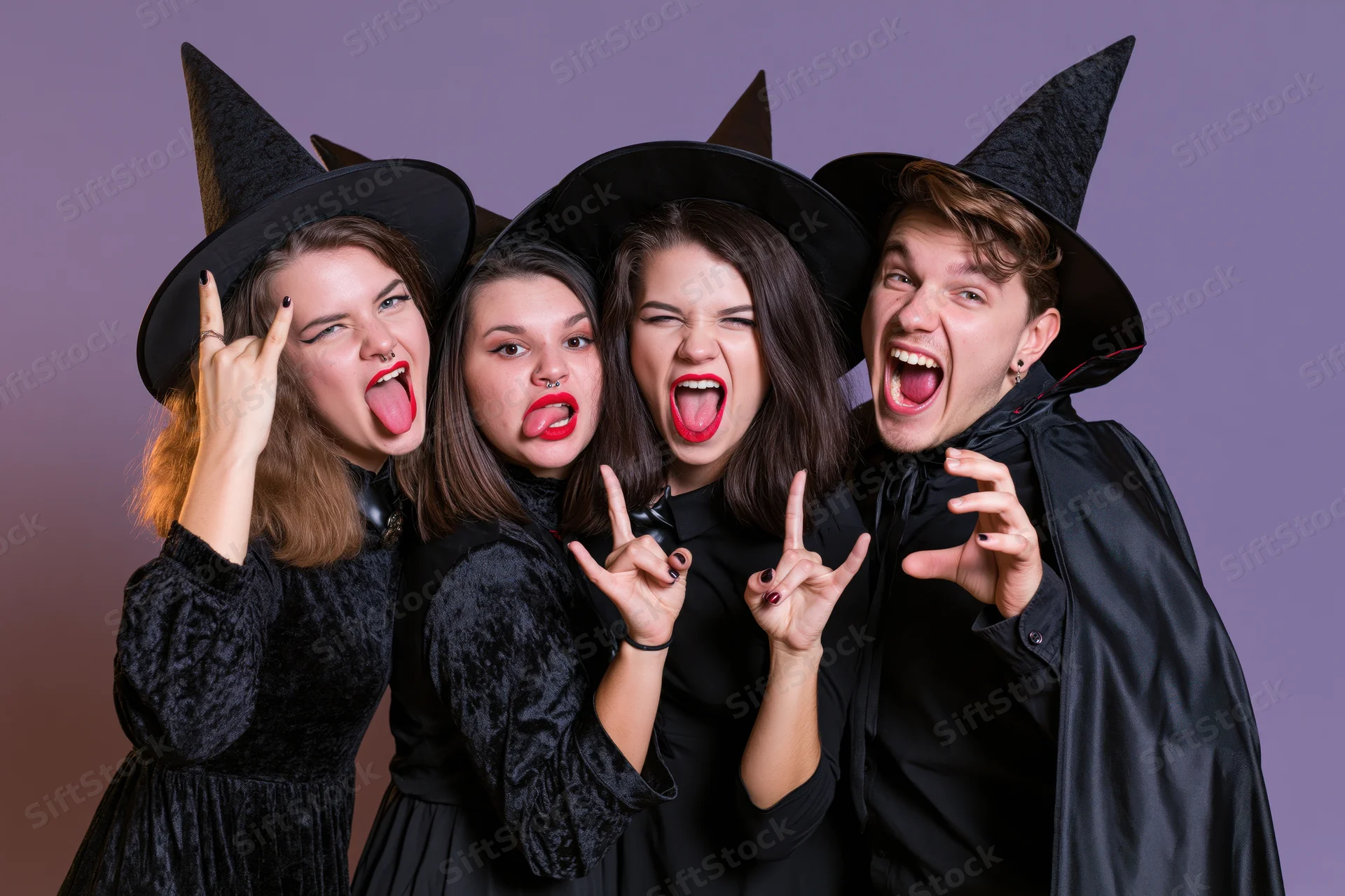 Four young adults in witch costumes posing playfully with tongues out and peace signs in a studio setting