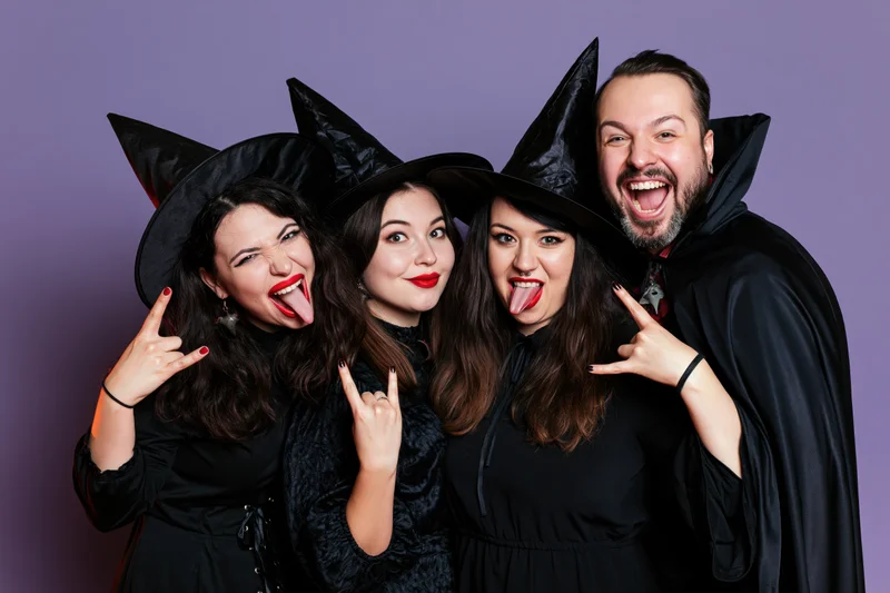 Four young adults in witch and vampire costumes posing playfully with rock hand gestures and tongues out against a purple studio background