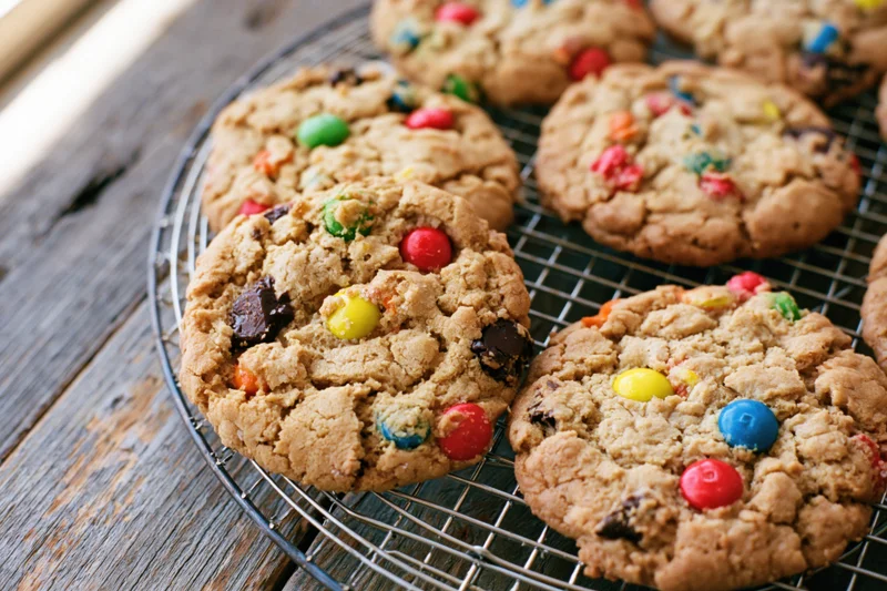 Freshly baked monster cookies with colorful candy and chocolate chips cooling on a wire rack on a wooden table