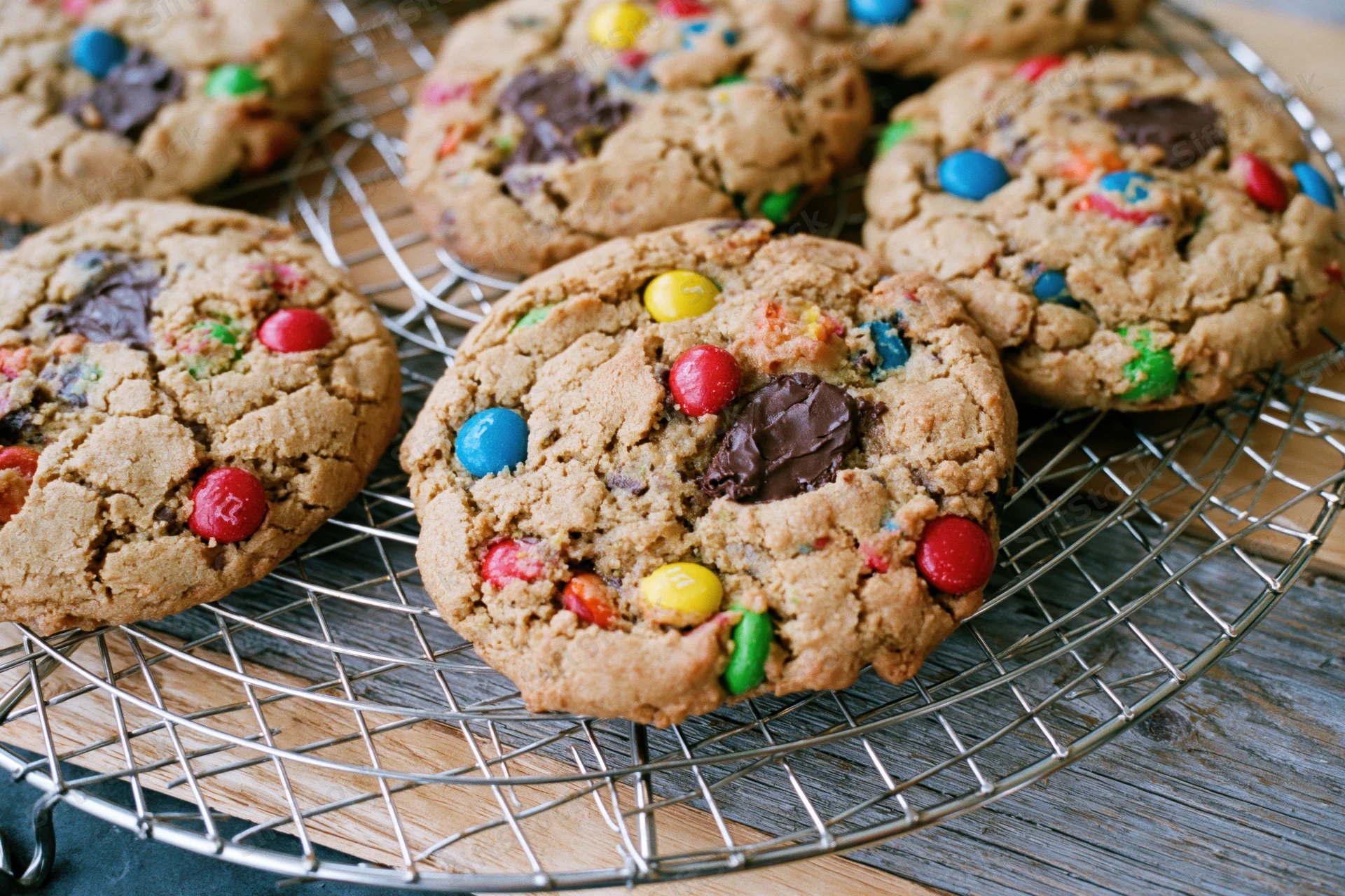 Freshly baked monster cookies with colorful candies and chocolate chunks cooling on a wire rack over a wooden surface