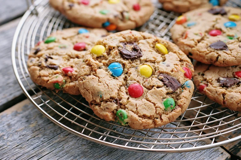 Freshly baked monster cookies with colorful candy-coated chocolates and chocolate chunks cooling on a wire rack on a wooden table