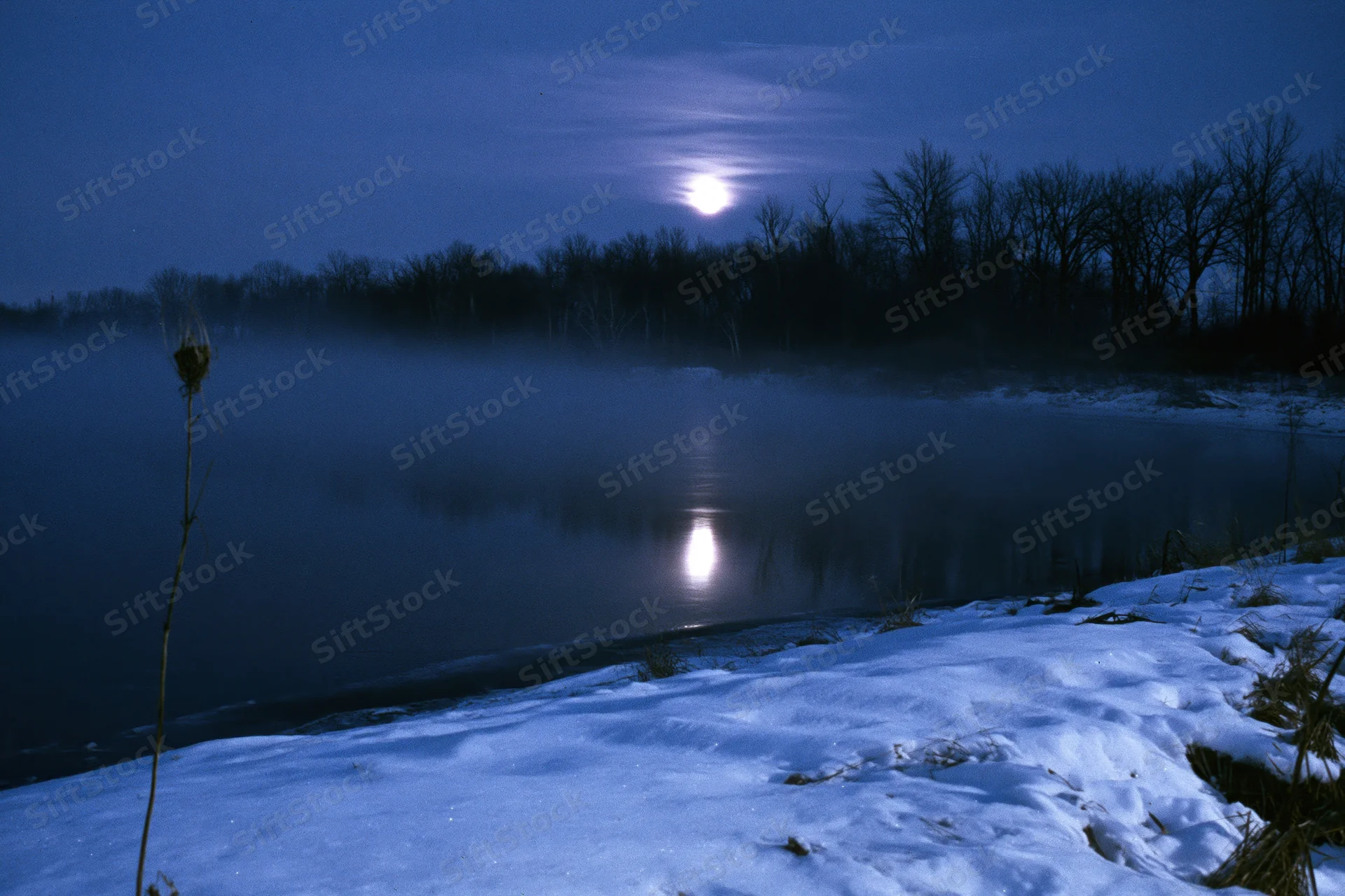 Full moon reflecting on a still winter river at night with snow-covered banks and mist rising from the water