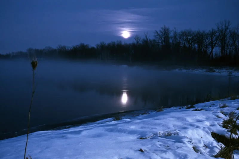 Full moon reflecting on a still winter river at night with snow-covered banks and mist rising from the water