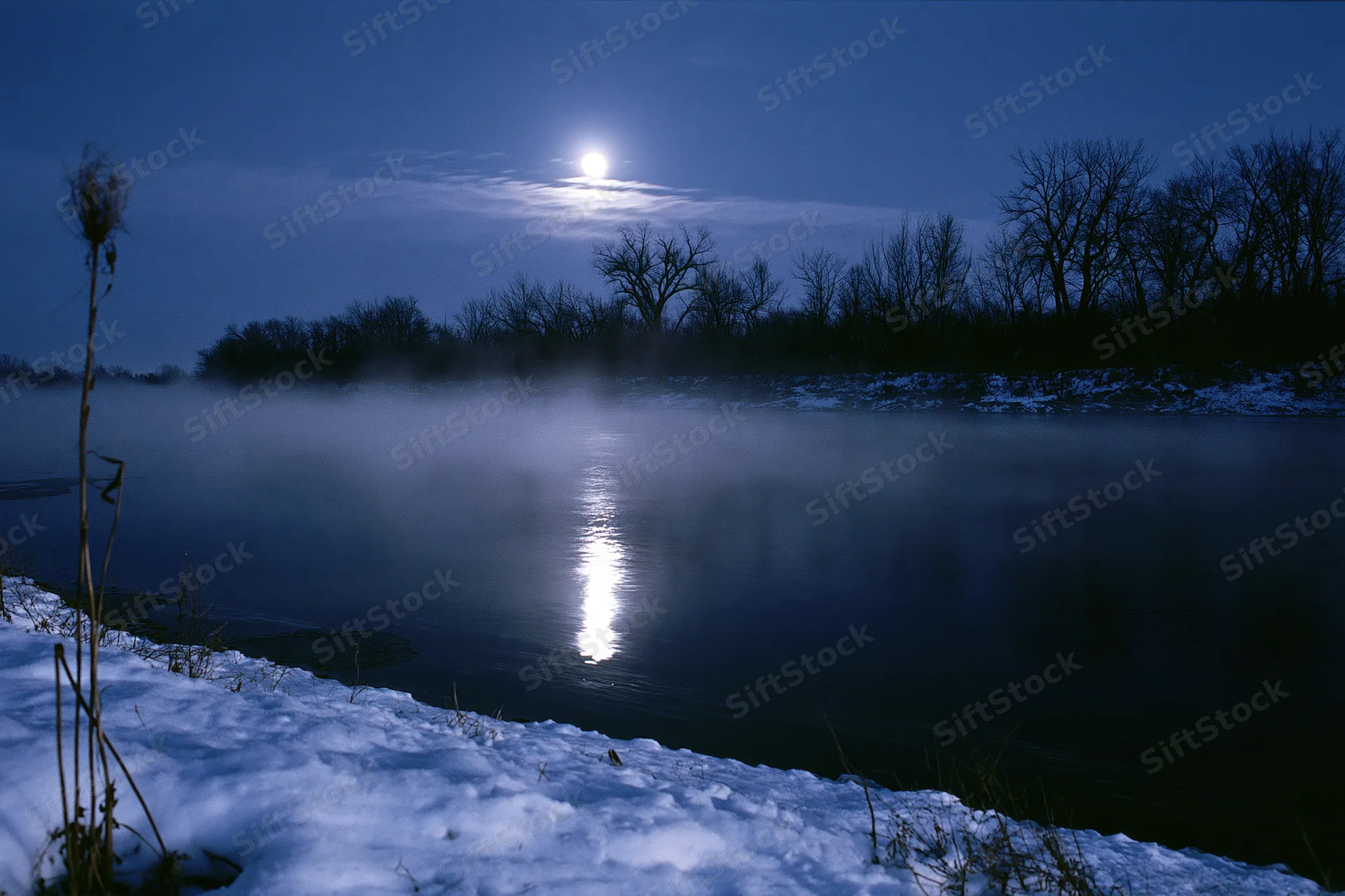 Full moon reflecting on a still winter river at night with mist rising from the water and snow along the bank